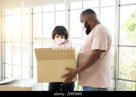 Mixed Race couple American African and Asian a surgical mask is carrying cardboard boxes in a new house on moving day. Concept of relocation, rental, Stock Photo