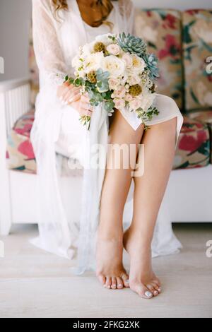 Barefoot bride in a lace robe sits on a sofa in a hotel room and holds a wedding bouquet in her hands, close-up Stock Photo