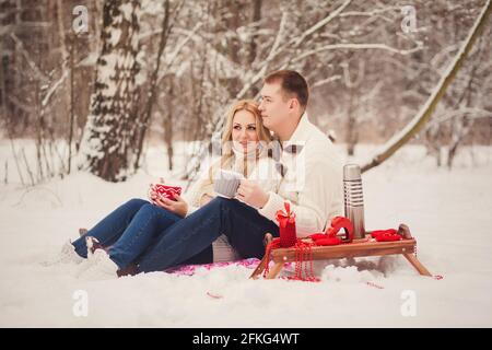 Young loving couple dressed in white sweater sitting with knitted coffee cups in the park Stock Photo