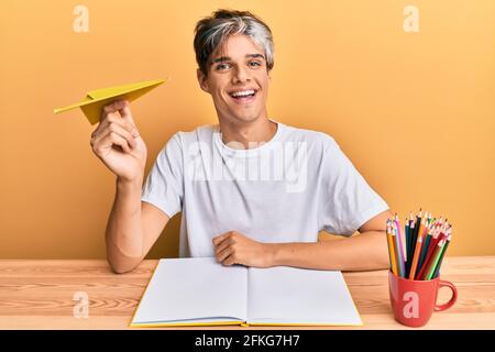 Young hispanic man studying and holding paper airplane sitting on the table looking positive and happy standing and smiling with a confident smile sho Stock Photo