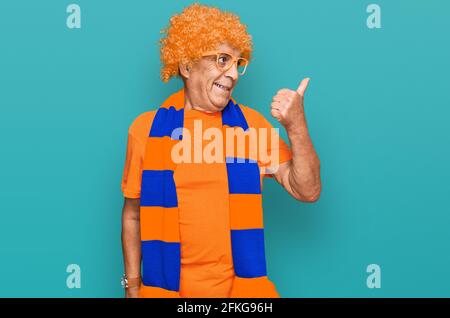 Senior hispanic man football hooligan cheering game smiling with happy face looking and pointing to the side with thumb up. Stock Photo