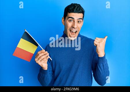 Handsome hispanic man holding belgium flag pointing thumb up to the side smiling happy with open mouth Stock Photo