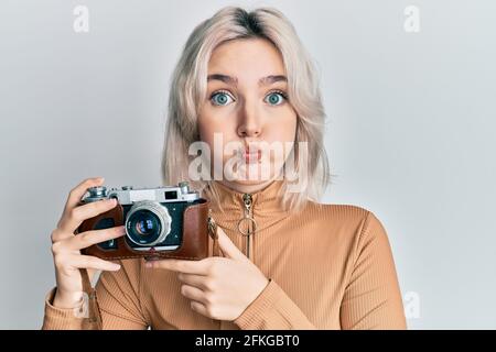 Young blonde girl holding vintage camera puffing cheeks with funny face. mouth inflated with air, catching air. Stock Photo