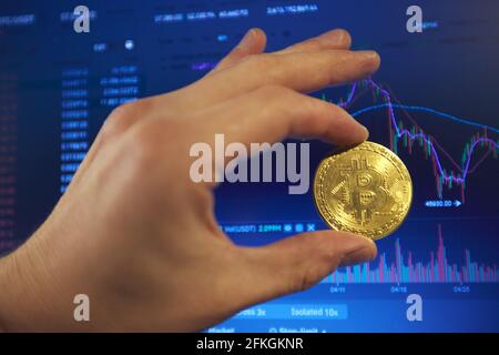 A man holds a gold bitcoin coin in his hand. The graphs of changes in cryptocurrency rates are shown on the laptop monitor Stock Photo