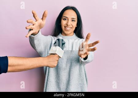 Young hispanic girl being interviewed by reporter holding microphone looking at the camera smiling with open arms for hug. cheerful expression embraci Stock Photo