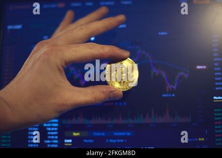 A man holds a gold bitcoin coin in his hand. The graphs of changes in cryptocurrency rates are shown on the laptop monitor Stock Photo