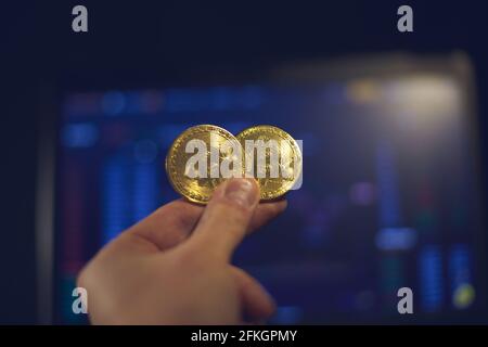 A man holds a gold bitcoin coin in his hand. The graphs of changes in cryptocurrency rates are shown on the laptop monitor Stock Photo
