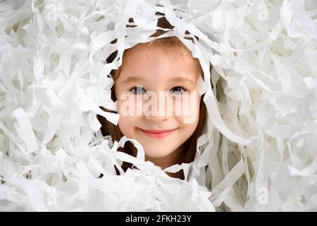 Kid looks out of hole in paper strips pile on party, portrait of cute little girl having fun with confetti. Adorable child face on white background, f Stock Photo