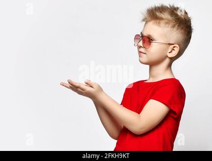 Portrait of blonde kid boy in red t-shirt and sunglasses standing holding hands up with open palms, holding blank space Stock Photo