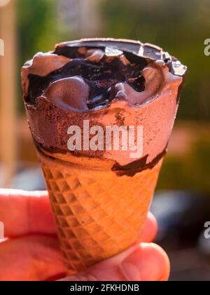 Chocolate ice cream cone held against a greenery and parked cars blurred in the background Stock Photo