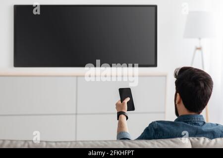 Young man watching television and using smart TV remote control application on smartphone, back view Stock Photo