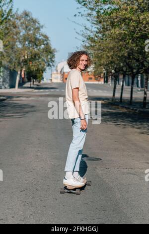 Caucasian red-haired man using a skateboard. He wears casual clothes. He skates down the road of a city surrounded by trees. Sunny summer day with har Stock Photo