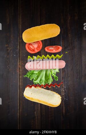 Levitating hot dog ingredients: sausage, mustard, ketchup, lettuce, tomatoes and bun on brown wooden plank surface. Stock Photo