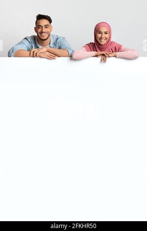 Nice Offer. Cheerful Muslim Spouses Leaning At Blank White Advertisement Board Stock Photo