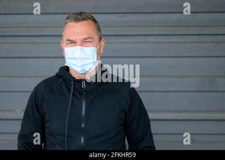 Middle-aged man wearing a face mask staring thoughtfully at camera with an intense expression as he poses outdoors against a grey wall during the Covi Stock Photo