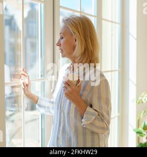 Enjoying new day. Side view of thoughtful beautiful mature woman holding cup of coffee or tea, looking in window and thinking or dreaming about something while spending free time in morning at home Stock Photo