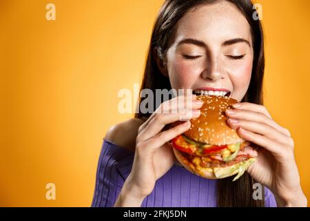 Girl bites cheeseburger with satisfaction. Woman eating delicious hambuger with pleasure and smile, have bite on lunch break at work, order takeaway Stock Photo