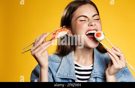 Happy girl eats sushi with satisfaction, holds sashimi and bites sushi roll with smile, order delicious takeaway in smartphone app, satisfied by fast Stock Photo