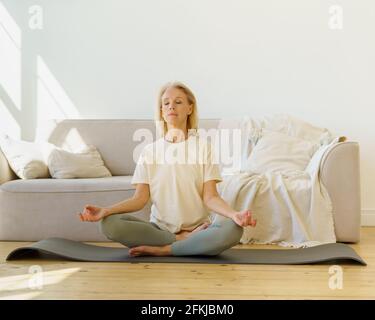 Peaceful senior woman in lotus position meditation with closed eyes at home while sitting on yoga mat on floor, full length. Calm elderly lady practicing meditation techniques and yoga indoors Stock Photo