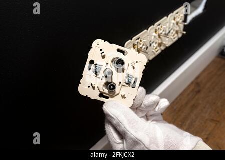An electrician in white gloves is installing a TV socket and satellite connector in the black wall of the room. Stock Photo