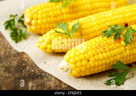 Three cobs of corn grilled on barbecue fire laid on dark stone table with parsley decoration. Healthy summer food concept. Clean eating habits. Backgr Stock Photo