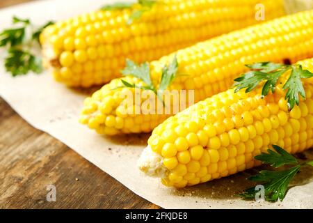 Three cobs of corn grilled on barbecue fire laid on dark stone table with parsley decoration. Healthy summer food concept. Clean eating habits. Backgr Stock Photo