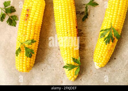 Three cobs of corn grilled on barbecue fire laid on dark stone table with parsley decoration. Healthy summer food concept. Clean eating habits. Backgr Stock Photo