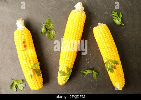 Three cobs of corn grilled on barbecue fire laid on dark stone table with parsley decoration. Healthy summer food concept. Clean eating habits. Backgr Stock Photo