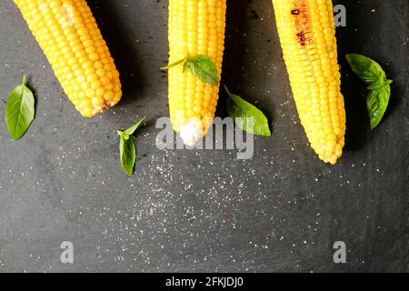 Three cobs of corn grilled on barbecue fire laid with green basil leaves and salt decoration on dark stone table. Clean eating habits concept. Backgro Stock Photo