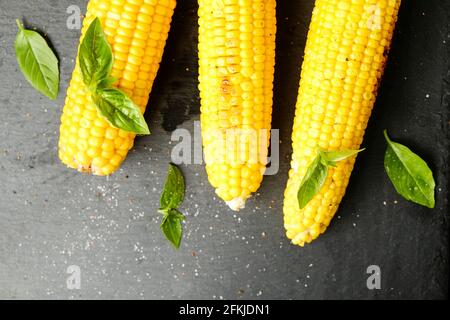 Three cobs of corn grilled on barbecue fire laid with green basil leaves and salt decoration on dark stone table. Clean eating habits concept. Backgro Stock Photo