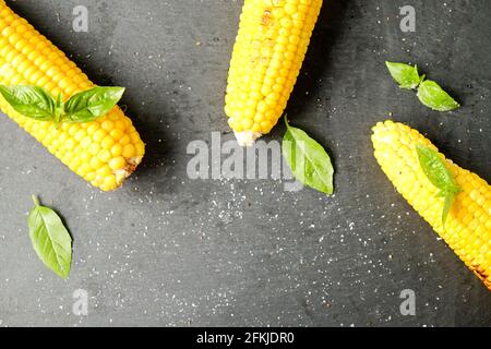 Three cobs of corn grilled on barbecue fire laid with green basil leaves and salt decoration on dark stone table. Clean eating habits concept. Backgro Stock Photo