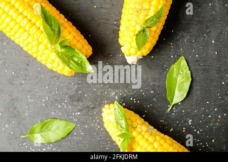 Three cobs of corn grilled on barbecue fire laid with green basil leaves and salt decoration on dark stone table. Clean eating habits concept. Backgro Stock Photo