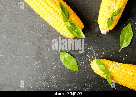 Three cobs of corn grilled on barbecue fire laid with green basil leaves and salt decoration on dark stone table. Clean eating habits concept. Backgro Stock Photo