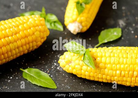 Three cobs of corn grilled on barbecue fire laid with green basil leaves and salt decoration on dark stone table. Clean eating habits concept. Backgro Stock Photo