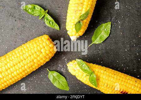 Three cobs of corn grilled on barbecue fire laid with green basil leaves and salt decoration on dark stone table. Clean eating habits concept. Backgro Stock Photo