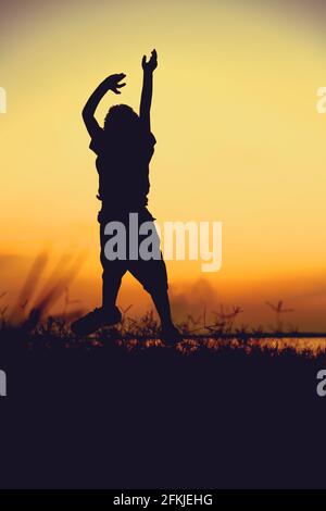 Silhouette back view of child jumping against sunset. Boy enjoying the view at riverside. Happy time on vacation. Vignette style. Stock Photo