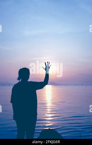 Silhouette back view of woman relax at seaside and showing stop gesture. The sun was hiding from hand on colorful blue sky background. Vintage picture Stock Photo