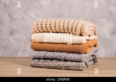 Bunch of knitted warm pastel color sweaters with different knitting patterns folded in stack on brown wooden table, grunged concrete wall background. Stock Photo