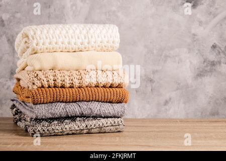 Bunch of knitted warm pastel color sweaters with different knitting patterns folded in stack on brown wooden table, grunged concrete wall background. Stock Photo