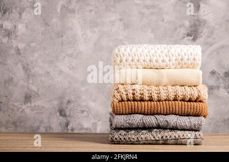 Bunch of knitted warm pastel color sweaters with different knitting patterns folded in stack on brown wooden table, grunged concrete wall background. Stock Photo