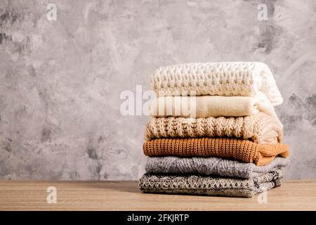 Bunch of knitted warm pastel color sweaters with different knitting patterns folded in stack on brown wooden table, grunged concrete wall background. Stock Photo