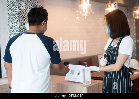 Asian waitress woman wearing face masks and holding an infrared thermometer and recommend customers to use hand alcohol sanitizer before entering the Stock Photo