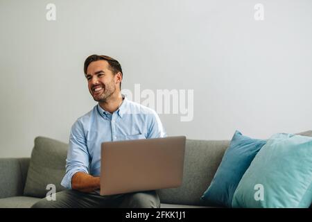Smiling man sitting on sofa in office working on laptop and looking away. Happy freelancer using laptop in lobby. Stock Photo