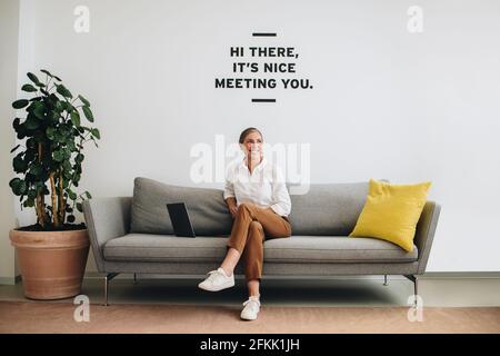 Mature woman sitting on sofa at office lobby. Senior freelance worker looking away and smiling. Stock Photo