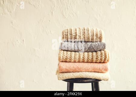 Bunch of knitted pastel color sweaters, different knitting patterns folded in neat stack on black wooden barstool, grunged texture wall background. Fa Stock Photo