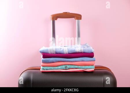 Stack of colorful perfectly folded clothing on plastic suitcase luggage. Pile of different pastel color shirts and sweaters on pale pink wall backgrou Stock Photo