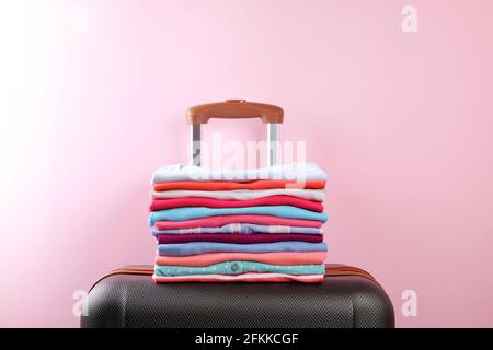 Stack of colorful perfectly folded clothing on plastic suitcase luggage. Pile of different pastel color shirts and sweaters on pale pink wall backgrou Stock Photo