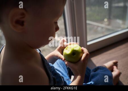 Child missing his mother concept. Little cute blond three year old boy sitting on windowsill touching window glass, looking sad. Stock Photo