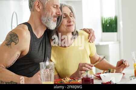 Happy mature husband embracing older wife enjoying having breakfast at home. Stock Photo
