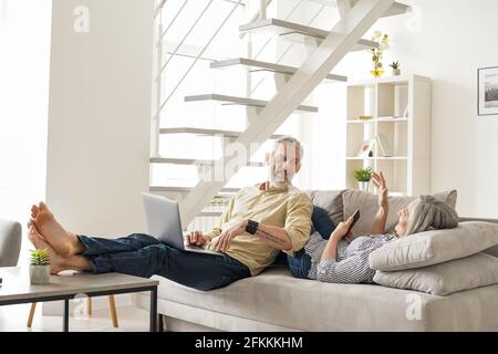 Happy mature older couple talking using devices relaxing at home on couch. Stock Photo